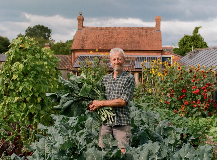 Charles Dowding standing in his no dig garden, holding freshly harvested vegetables, surrounded by vibrant greenery and flowers with a red brick house in the background.