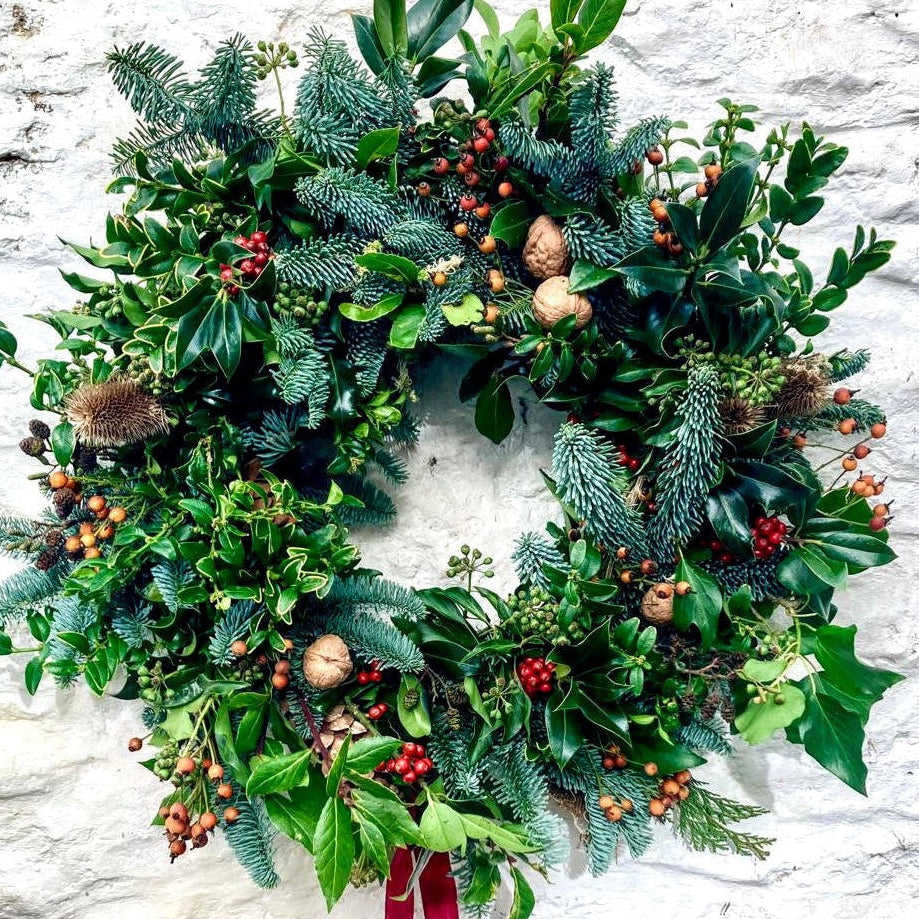 A festive green Christmas wreath with pine branches, holly leaves, red berries, acorns, and a long dark red ribbon hanging from the bottom, displayed against a white textured wall.