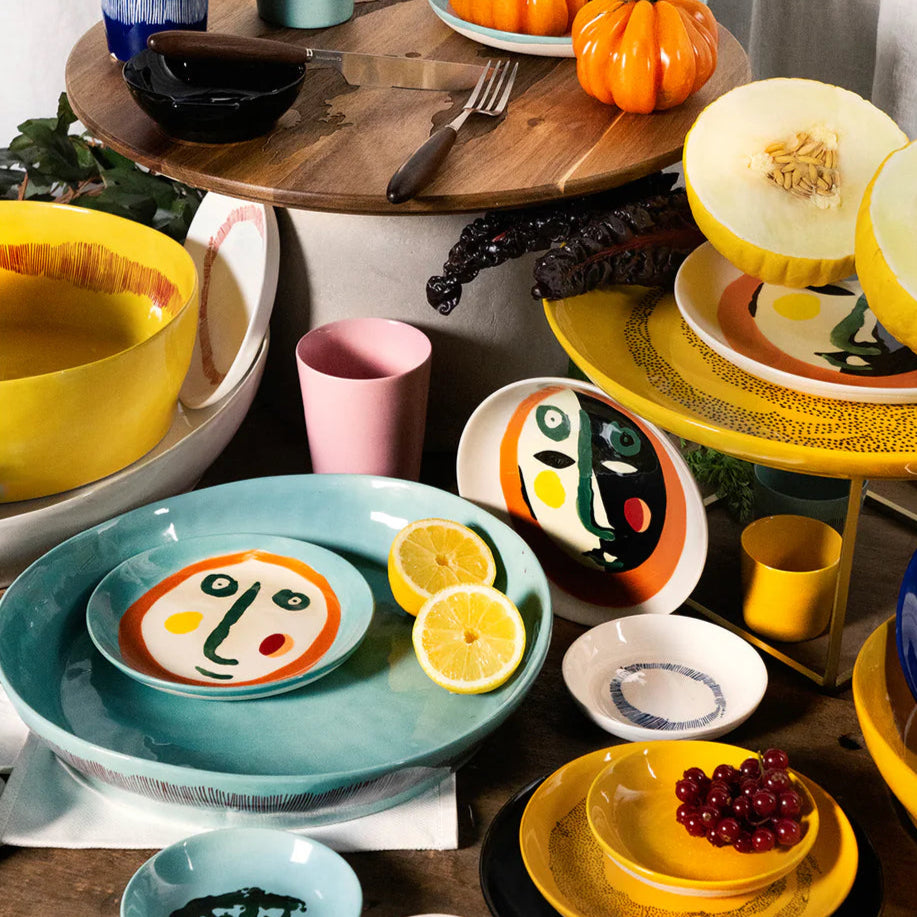 A series of colourful plates laid out on a wooden table with fruit, such as melon, tomatoes and grapes