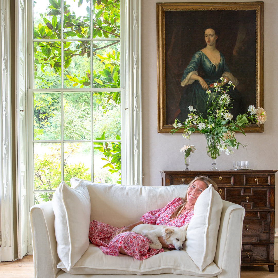 Sophie lying on a white sofa with a dog, surrounded by decor including a painting and flowers.