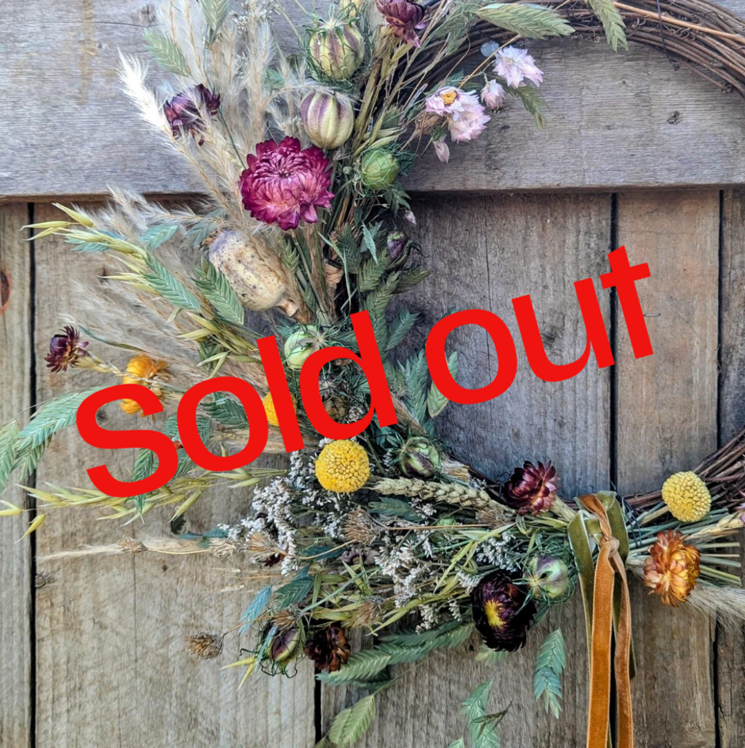 A handcrafted autumnal wreath made with dried flowers, seed heads, grasses and foliage in warm, natural tones, resting against rustic wooden boards. Sold out text over top.
